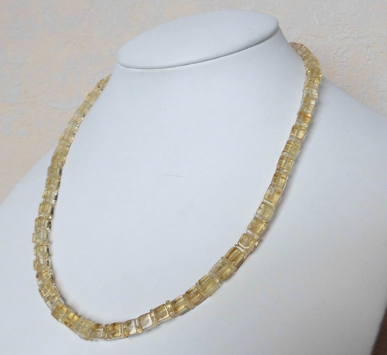 May include: A gold necklace made of square-shaped beads. The necklace is on a white mannequin.