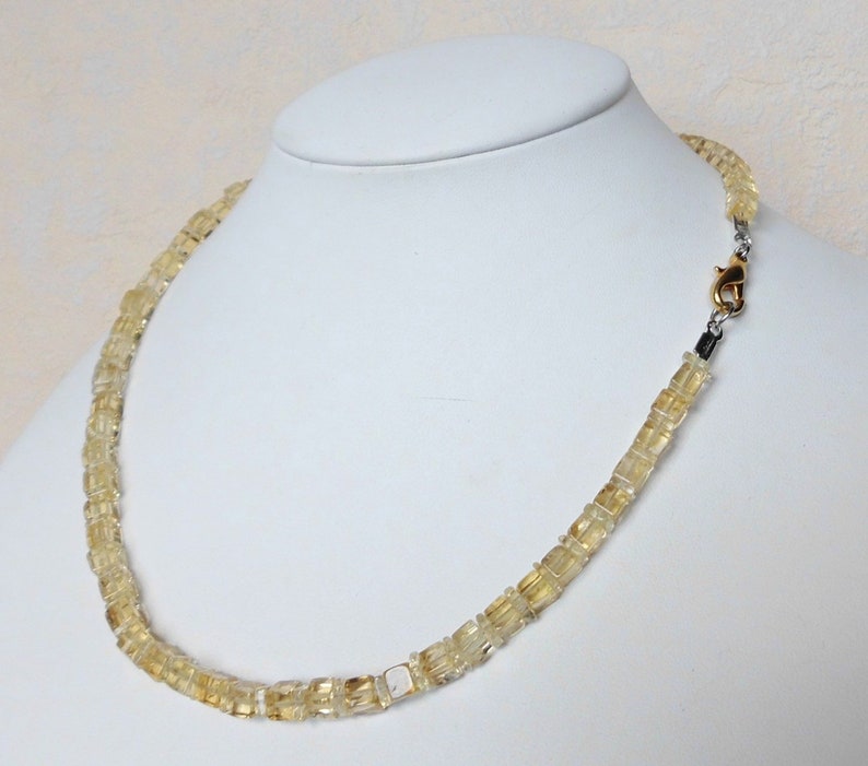 May include: A gold and clear citrine gemstone necklace with a gold clasp. The necklace is made of square-shaped beads.