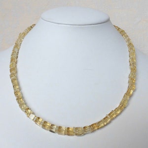 May include: A necklace made of light yellow, square-shaped beads. The necklace is on a white mannequin bust.