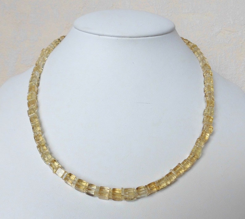 May include: A necklace made of light yellow, square-shaped beads. The necklace is on a white mannequin bust.