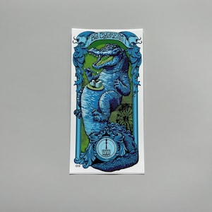 May include: A blue alligator with a clock face on its belly, holding a box with a clock face on it. The alligator is surrounded by a green and blue frame with the text "Big Cypress" and "The Gator Watch" in a banner.