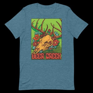 Puede incluir: Una camiseta azul turquesa con un estampado gráfico de una calavera de ciervo con cuernos rodeada de flores rojas y azules. El texto "Deer Creek" está impreso en letras rojas y naranjas debajo de la calavera.