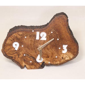 May include: A distinctive wooden clock made from a tree trunk slice. The clock face displays white numerals at 3, 9, and 12 o'clock, with white dots for other hours. The clock hands are gold-coloured. The clock measures approximately 25 cm in diameter.