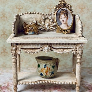 May include: A miniature cream-coloured display table with a gold crown, a framed portrait, and a decorative ceramic pitcher. The table has ornate detailing and is set against a floral background. The table is reminiscent of antique furniture.