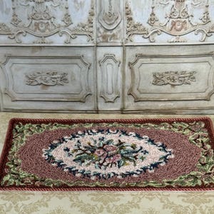 Könnte beinhalten: Ein rechteckiger Teppich mit floralem Design. Der Teppich hat ein zentrales Oval mit rosa und blauen Blumen auf weißem Grund, umgeben von einem rosa Feld und einem grünen Rand. Maße: ca. 30 cm x 15 cm.
