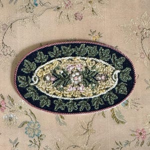 Könnte beinhalten: Ovales, handgenähtes Textil mit dunkelblauem Rand und einem zentralen Design aus Blumen und Blättern. Der Innenraum hat einen cremefarbenen Hintergrund mit rosa und grünen floralen Akzenten. Der Rand ist mit einer rosa Seilborte versehen.