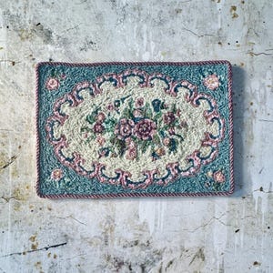 Könnte beinhalten: Ein rechteckiger, handgenähter Teppich mit floralem Design. Der Teppich hat ein ovales Zentrum aus cremefarbenem Stoff mit einem Bouquet aus rosa und grünen Blumen. Der Rand ist türkis mit rosa Schnörkeln und einem rosa Seilrand.
