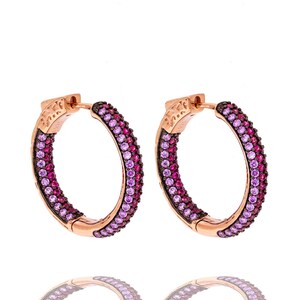 Peut inclure: Une paire de boucles d'oreilles créoles en or rose. Les créoles sont incrustées de pierres précieuses roses et violettes en dégradé. Les boucles d'oreilles sont posées sur un fond blanc, avec un léger reflet en dessous.