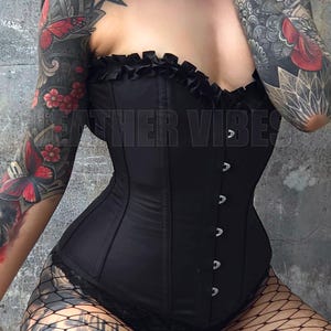 May include: A black corset with a ruffled top and silver buttons. The corset is worn over a black lace top and fishnet stockings. The person wearing the corset has tattoos on both arms.
