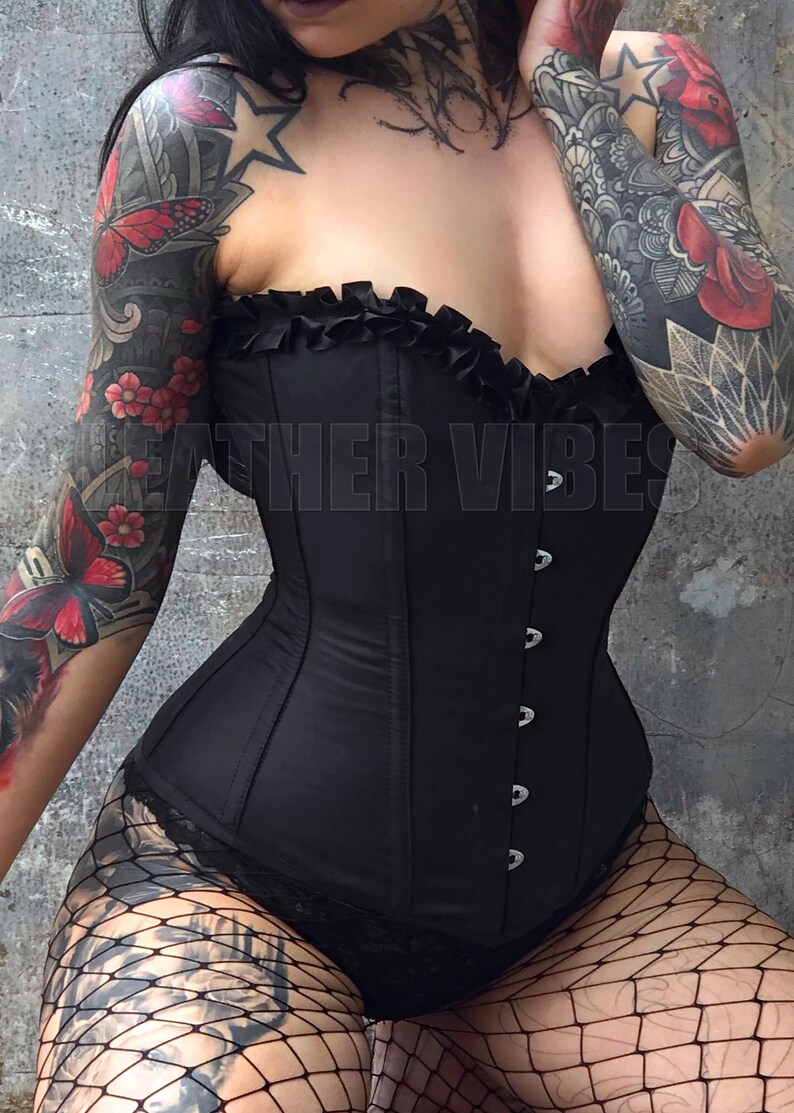 May include: A black corset with a ruffled top and silver buttons. The corset is worn over a black lace top and fishnet stockings. The person wearing the corset has tattoos on both arms.