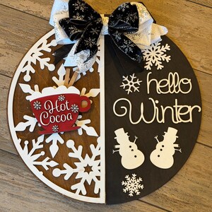 Op de afbeelding: Een ronde houten winterbord, in twee&euml;n verdeeld met een sneeuwvlokontwerp en de woorden "hello Winter". Voorzien van een rode "Hot Cocoa" mok en twee sneeuwpopfiguren. Bovenop een zwart-witte strik.