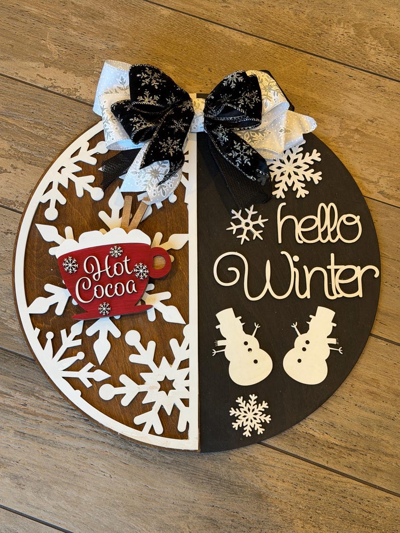 Op de afbeelding: Een ronde houten winterbord, in twee&euml;n verdeeld met een sneeuwvlokontwerp en de woorden "hello Winter". Voorzien van een rode "Hot Cocoa" mok en twee sneeuwpopfiguren. Bovenop een zwart-witte strik.