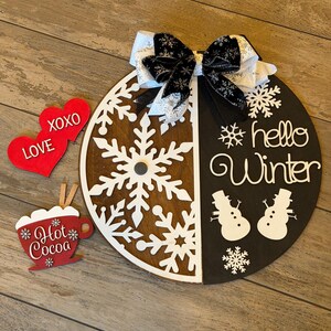 Op de afbeelding: Een ronde houten winterdecoratie met een sneeuwvlokontwerp en de tekst "hello Winter". Inclusief een decoratieve strik, twee rode hartvormige ornamenten met "XOXO LOVE" en een "Hot Cocoa" ornament. De diameter is ongeveer 30 cm.