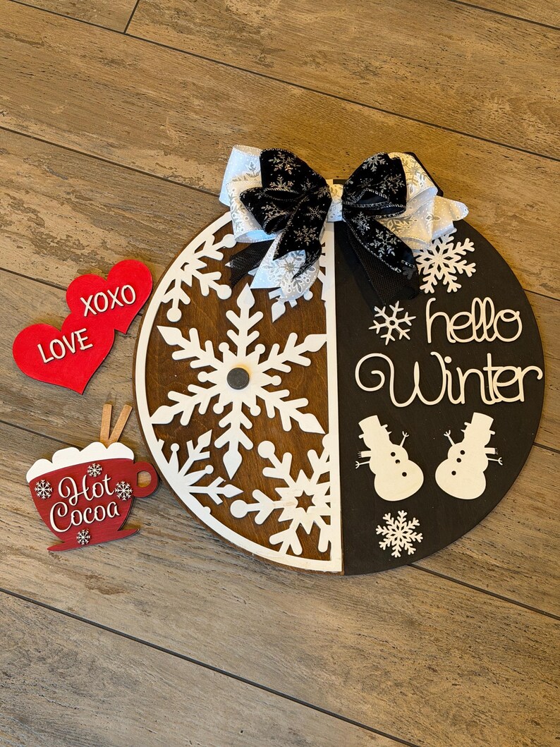 Op de afbeelding: Een ronde houten winterdecoratie met een sneeuwvlokontwerp en de tekst "hello Winter". Inclusief een decoratieve strik, twee rode hartvormige ornamenten met "XOXO LOVE" en een "Hot Cocoa" ornament. De diameter is ongeveer 30 cm.