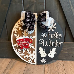Op de afbeelding: Een ronde houten winterbord, in twee&euml;n gesplitst met een sneeuwvlokontwerp en de woorden "hello Winter". Het bord heeft een rode "Hot Cocoa" mok en een zwart-witte strik.