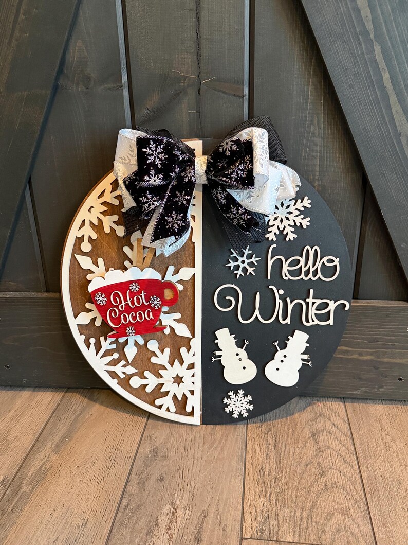 Op de afbeelding: Een ronde houten winterbord, in twee&euml;n gesplitst met een sneeuwvlokontwerp en de woorden "hello Winter". Het bord heeft een rode "Hot Cocoa" mok en een zwart-witte strik.