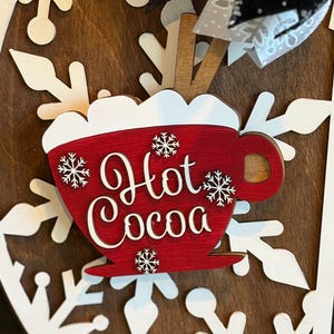 Op de afbeelding: Een houten bord met een rood "Hot Cocoa" mokontwerp, versierd met witte sneeuwvlokken. Het bord staat tegen een donkerbruine achtergrond en heeft witte sneeuwvlok uitsparingen. Een decoratieve strik is zichtbaar aan de bovenkant.