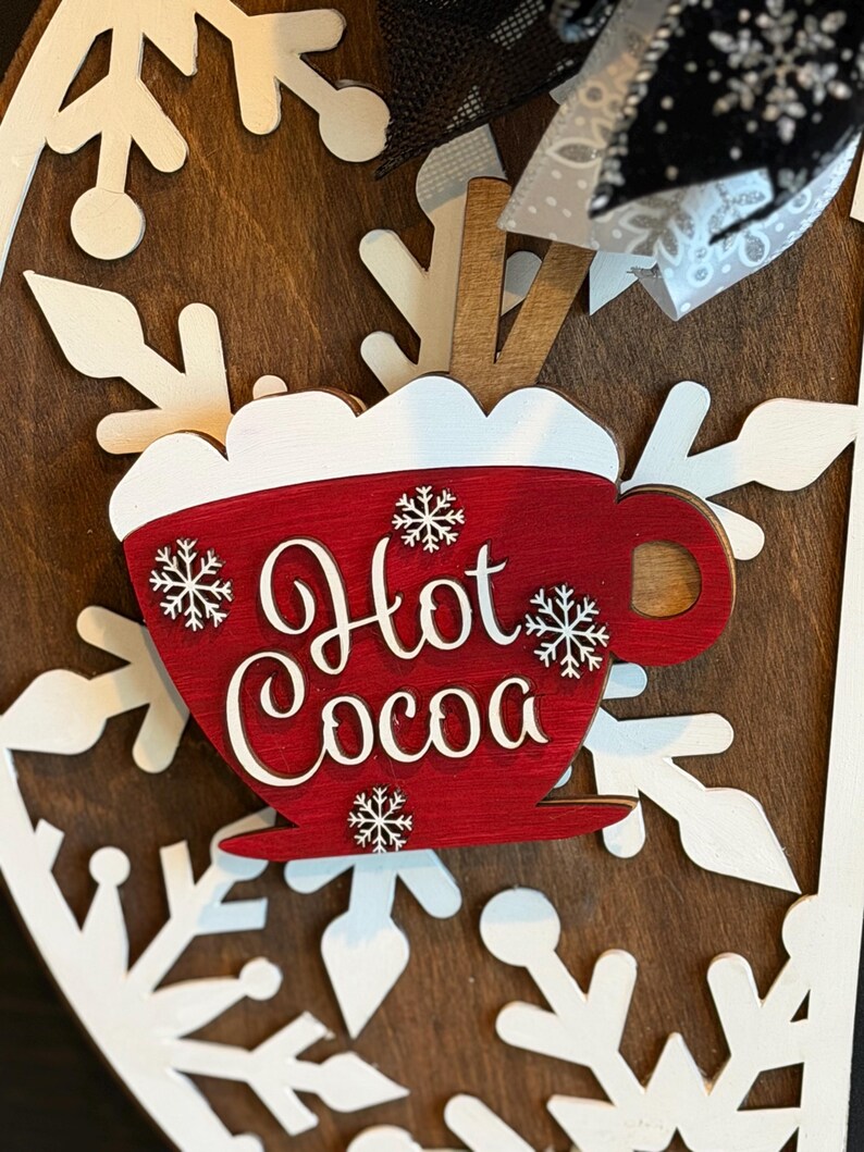 Op de afbeelding: Een houten bord met een rood "Hot Cocoa" mokontwerp, versierd met witte sneeuwvlokken. Het bord staat tegen een donkerbruine achtergrond en heeft witte sneeuwvlok uitsparingen. Een decoratieve strik is zichtbaar aan de bovenkant.