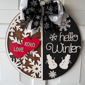 Op de afbeelding: Een ronde houten winterdeurhanger, verdeeld in bruine en zwarte helften. De hanger heeft sneeuwvlokken, sneeuwpoppen en de woorden "hello Winter". Twee rode harten met "LOVE XOXO" staan links, en een zwart-witte strik staat bovenaan.