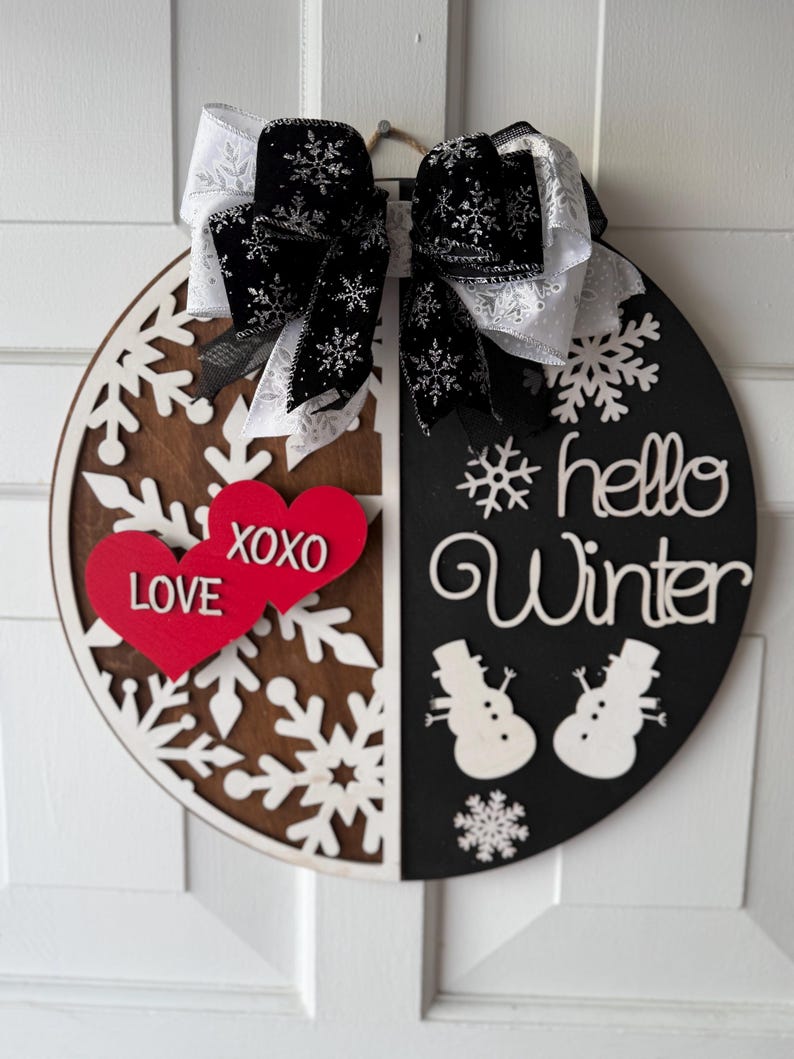 Op de afbeelding: Een ronde houten winterdeurhanger, verdeeld in bruine en zwarte helften. De hanger heeft sneeuwvlokken, sneeuwpoppen en de woorden "hello Winter". Twee rode harten met "LOVE XOXO" staan links, en een zwart-witte strik staat bovenaan.
