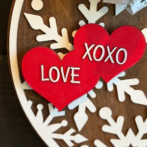 Op de afbeelding: Een ronde houten decoratie met een bruine achtergrond. Witte sneeuwvlokpatronen omlijsten twee rode harten. E&eacute;n hart toont "LOVE", de andere "XOXO", beide in het wit. Een zwart-witte strik is rechtsboven zichtbaar.