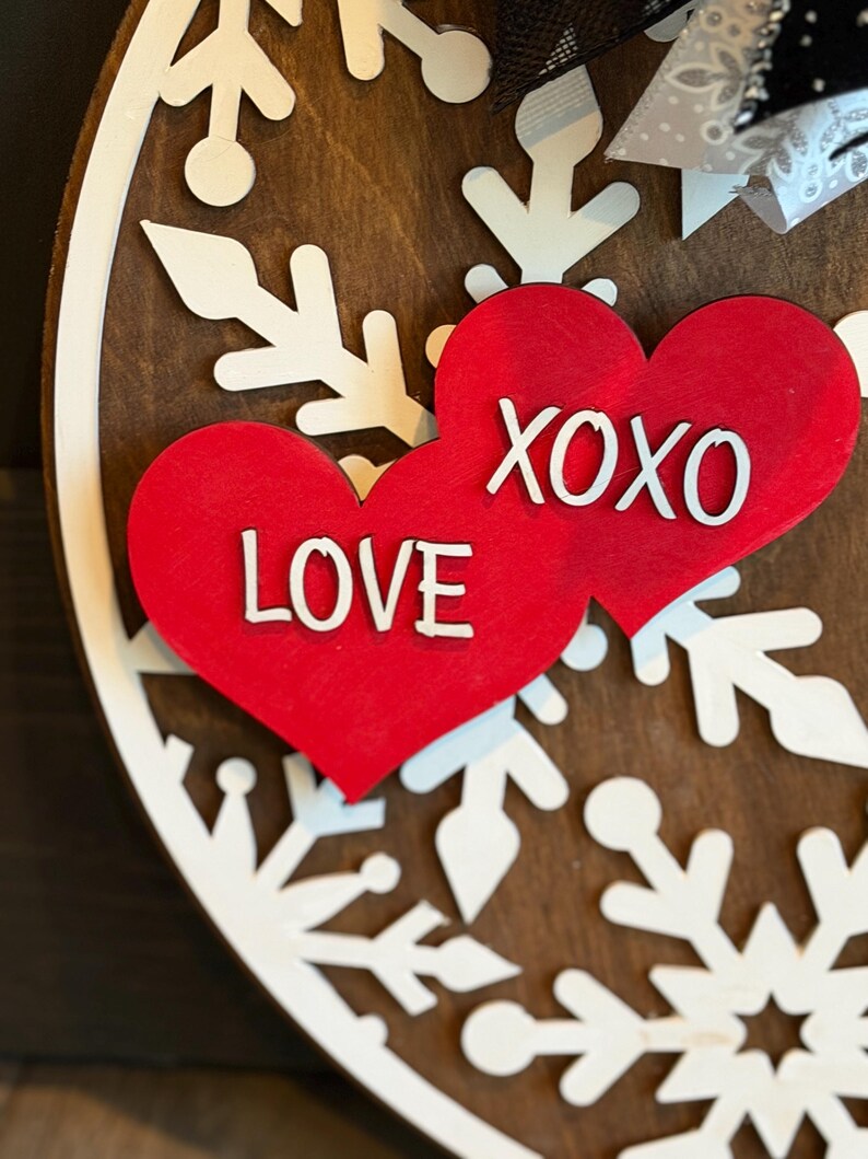 Op de afbeelding: Een ronde houten decoratie met een bruine achtergrond. Witte sneeuwvlokpatronen omlijsten twee rode harten. E&eacute;n hart toont "LOVE", de andere "XOXO", beide in het wit. Een zwart-witte strik is rechtsboven zichtbaar.