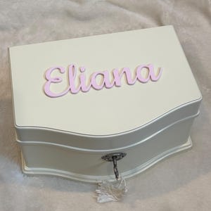 Puede incluir: Joyero color crema con el nombre "Eliana" en escritura rosa en la tapa. La caja tiene un diseño curvo y un ojo de cerradura con una borla. Un artículo de almacenamiento decorativo.