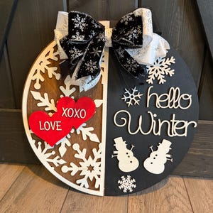 Op de afbeelding: Een ronde houten winterbord met een zwart-witte strik. Het bord is in twee&euml;n verdeeld, met aan de ene kant sneeuwvlokken en rode harten met "LOVE XOXO". De andere kant zegt "hello Winter" met sneeuwpopuitsparingen.