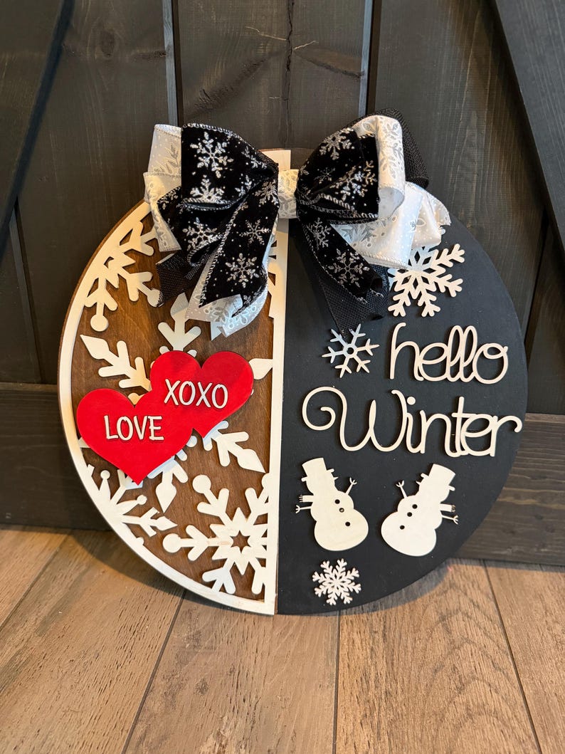 Op de afbeelding: Een ronde houten winterbord met een zwart-witte strik. Het bord is in twee&euml;n verdeeld, met aan de ene kant sneeuwvlokken en rode harten met "LOVE XOXO". De andere kant zegt "hello Winter" met sneeuwpopuitsparingen.