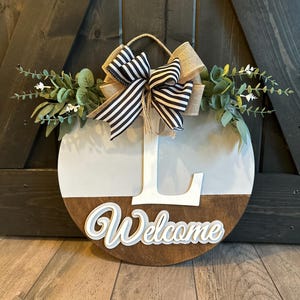 May include: Round wooden welcome sign with a white upper section and a brown lower section. The word "Welcome" is in white script. A large white letter "L" is centered above the word. The sign is decorated with greenery and a black and white striped bow.