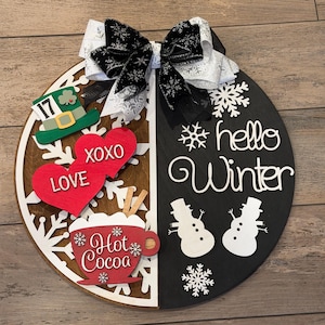 Op de afbeelding: Een ronde houten plank, verdeeld in bruine en zwarte helften, versierd met een decoratieve strik. De bruine kant toont een groene hoed, rode harten met "LOVE" en "XOXO", en een "Hot Cocoa" beker. De zwarte kant zegt "hello Winter" met sneeuwmannen.
