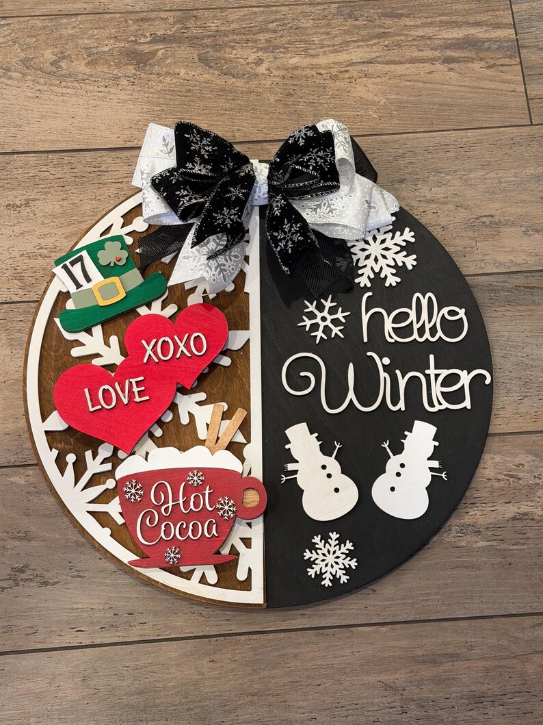 Op de afbeelding: Een ronde houten plank, verdeeld in bruine en zwarte helften, versierd met een decoratieve strik. De bruine kant toont een groene hoed, rode harten met "LOVE" en "XOXO", en een "Hot Cocoa" beker. De zwarte kant zegt "hello Winter" met sneeuwmannen.