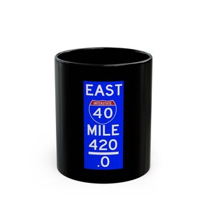 May include: A black ceramic mug featuring a blue and white road sign design. The sign reads "EAST INTERSTATE 40 MILE 420 .0". The mug is cylindrical and has a glossy finish.