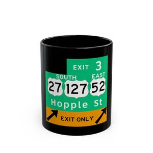 May include: Black ceramic mug featuring a green and white highway exit sign design. The sign indicates Exit 3, with route numbers 27, 127, and 52, and the street name Hopple St. The mug has a black handle and interior.