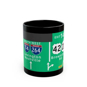 May include: Black coffee mug with a green wrap-around design featuring highway signs. The signs indicate Interstate 74 and 264, with directions to Lexington and Nashville. The mug also shows Exit 14 and Brownsb Rd.