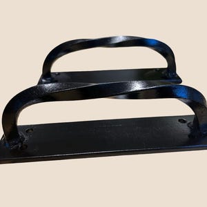 May include: Two black, wrought iron door pulls. Each pull features a rectangular base with a curved, twisted handle. The handles are arched and have a decorative, textured design. The hardware is painted black.