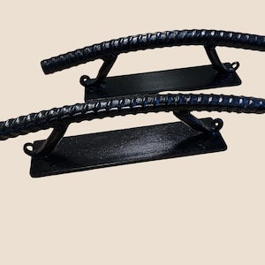 May include: Two black metal handles with a textured, rebar-like design. Each handle is mounted on a rectangular base with curved supports. The handles are designed for a door or cabinet.