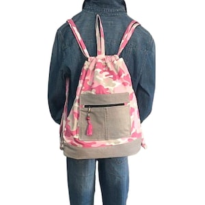 May include: Pink and gray camouflage drawstring backpack with a gray denim pocket and a pink tassel. The backpack has pink straps and a gray bottom. The bag is worn over a denim jacket.