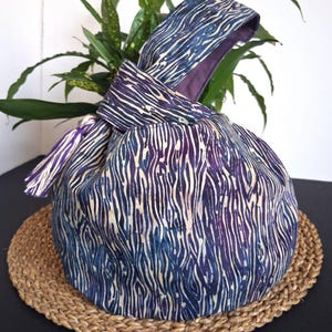 May include: A round, fabric bag with a unique pattern of white, blue, and purple vertical lines. The bag is tied at the top with a matching fabric knot and a purple tassel. It sits on a woven, circular mat.
