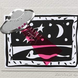 May include: A white card with black ink and a silver sticker of a flying saucer with pink rhinestones. The card has a black and white illustration of a night sky with a crescent moon and stars. The text "Thinking of you" is written in black ink.