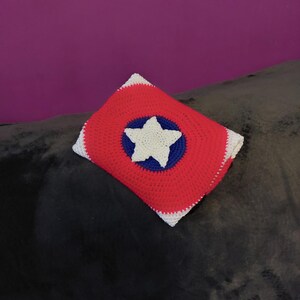 May include: A crocheted pillow with a red background, a blue circle, and a white star in the center. The pillow has a white border and is placed on a dark gray surface. The pillow's design is reminiscent of a shield.