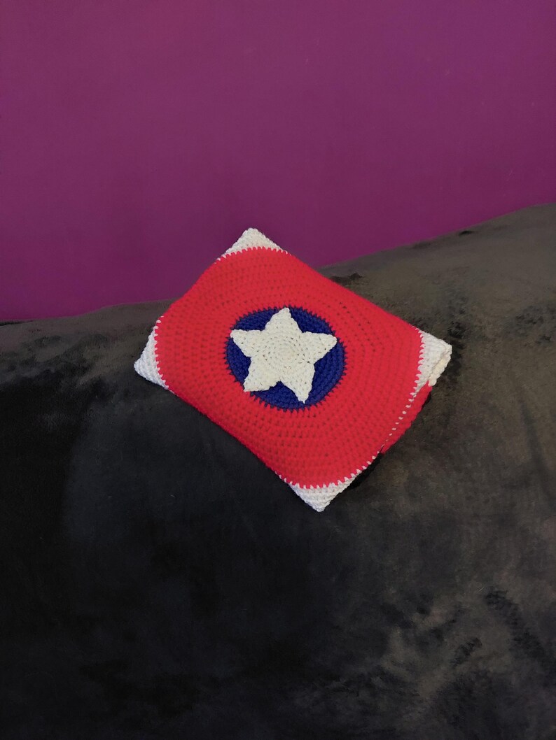 May include: A crocheted pillow with a red background, a blue circle, and a white star in the center. The pillow has a white border and is placed on a dark gray surface. The pillow's design is reminiscent of a shield.