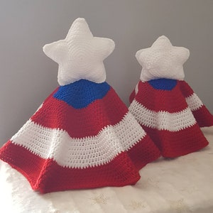 May include: Two handmade crochet star-shaped toppers with red, white, and blue striped capes. The white stars sit atop the capes, which are reminiscent of a flag. The items are likely decorative, possibly for a holiday or patriotic theme.