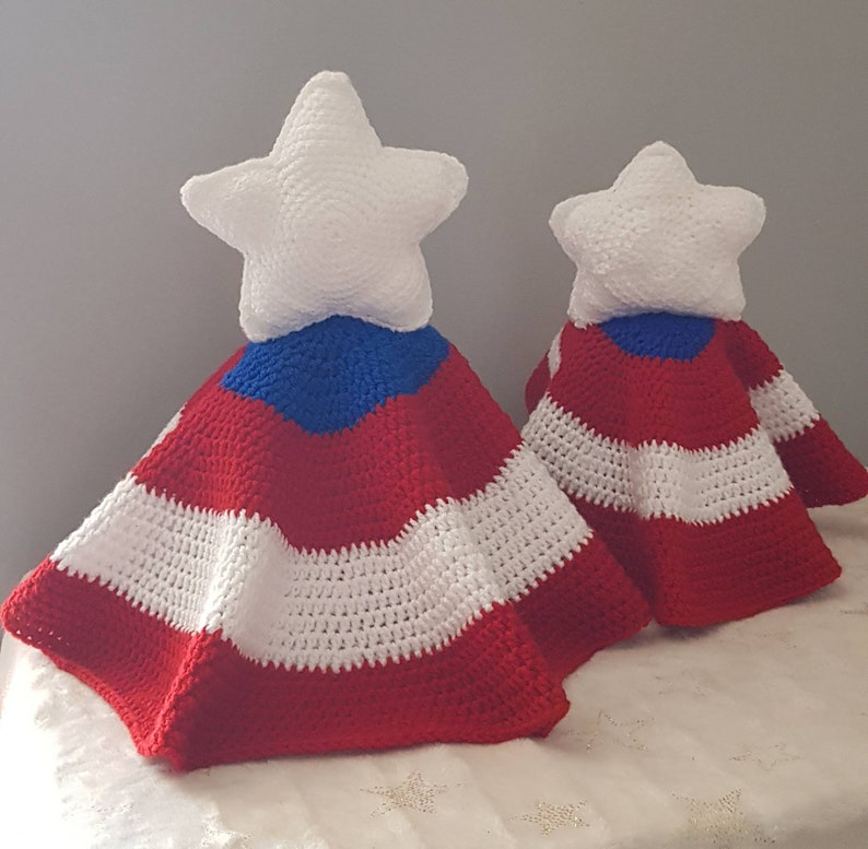 May include: Two handmade crochet star-shaped toppers with red, white, and blue striped capes. The white stars sit atop the capes, which are reminiscent of a flag. The items are likely decorative, possibly for a holiday or patriotic theme.