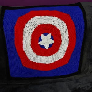 May include: A handmade crochet blanket featuring a shield design in red, white, and blue. The blanket has a white star in the center, surrounded by concentric circles of red, white, and blue. The edges are black.
