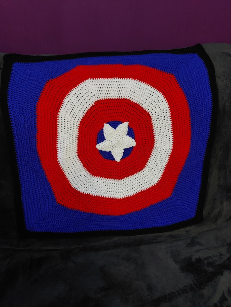 May include: A handmade crochet blanket featuring a shield design in red, white, and blue. The blanket has a white star in the center, surrounded by concentric circles of red, white, and blue. The edges are black.