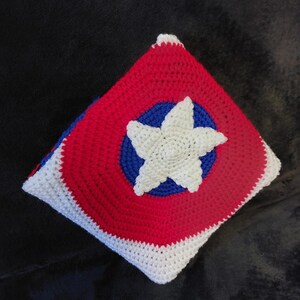 May include: A handmade crochet pillow with a patriotic design. The square pillow features a red background with a blue circle and a white star in the center. The edges are white, and the pillow is likely filled with soft material.