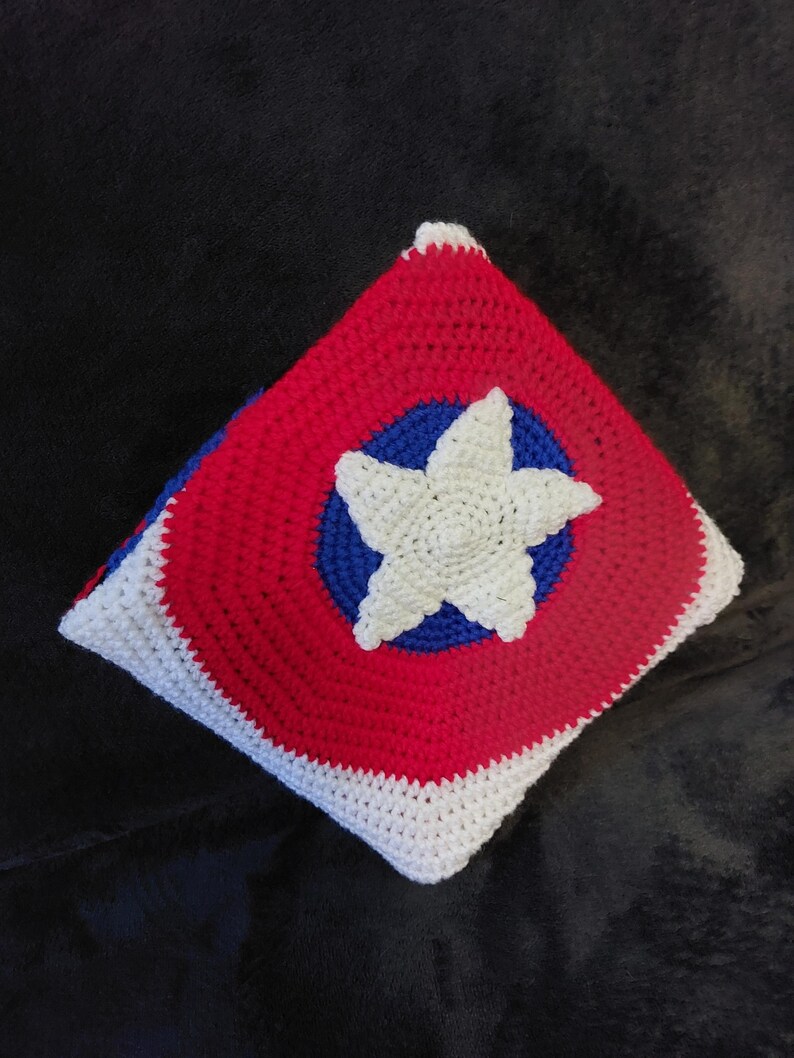 May include: A handmade crochet pillow with a patriotic design. The square pillow features a red background with a blue circle and a white star in the center. The edges are white, and the pillow is likely filled with soft material.