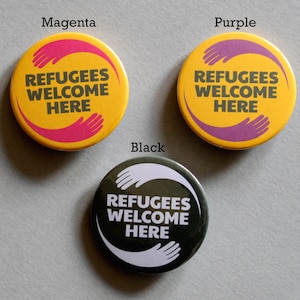 May include: Three round buttons with the text "Refugees Welcome Here" in black lettering on a yellow background. The buttons have different coloured handprints around the text: magenta, purple, and black.