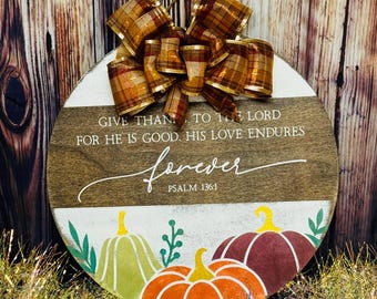 May include: A round wooden sign with a white background and a brown border. The sign features a fall-themed design with pumpkins and leaves. The text "GIVE THANKS TO THE LORD FOR HE IS GOOD. HIS LOVE ENDURES FOREVER PSALM 136:1" is written in black on the sign. A brown and gold plaid bow is attached to the top of the sign.